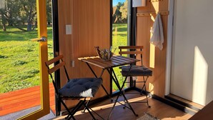 A room with a wooden door, a small table with a bowl and magazines, two chairs, and a view of a green outdoor area. - Tiny Home in the Hills by Tiny Away (Forrestfield)