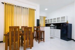 A modern kitchen with white cabinets, a black refrigerator, and a dining area with wooden chairs and yellow curtains. - Skyport Villa Katunayaka (Katunayake)
