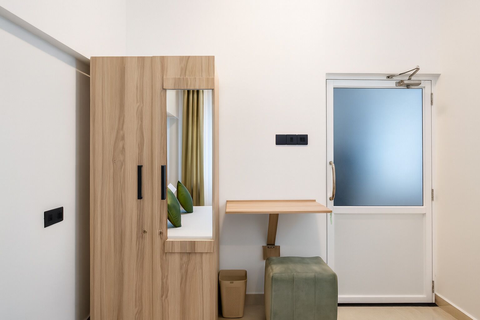 Una habitación con un armario de madera, una mesita pequeña, un banco, una puerta con cristal esmerilado y un interruptor de luz en la pared.
