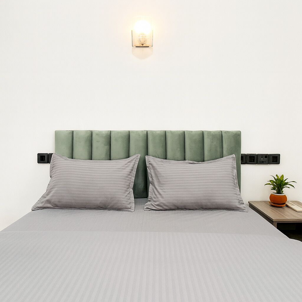 A bed with a green upholstered headboard, two grey pillows, a grey striped bedspread, a bedside table with a plant, and a wall-mounted light fixture.