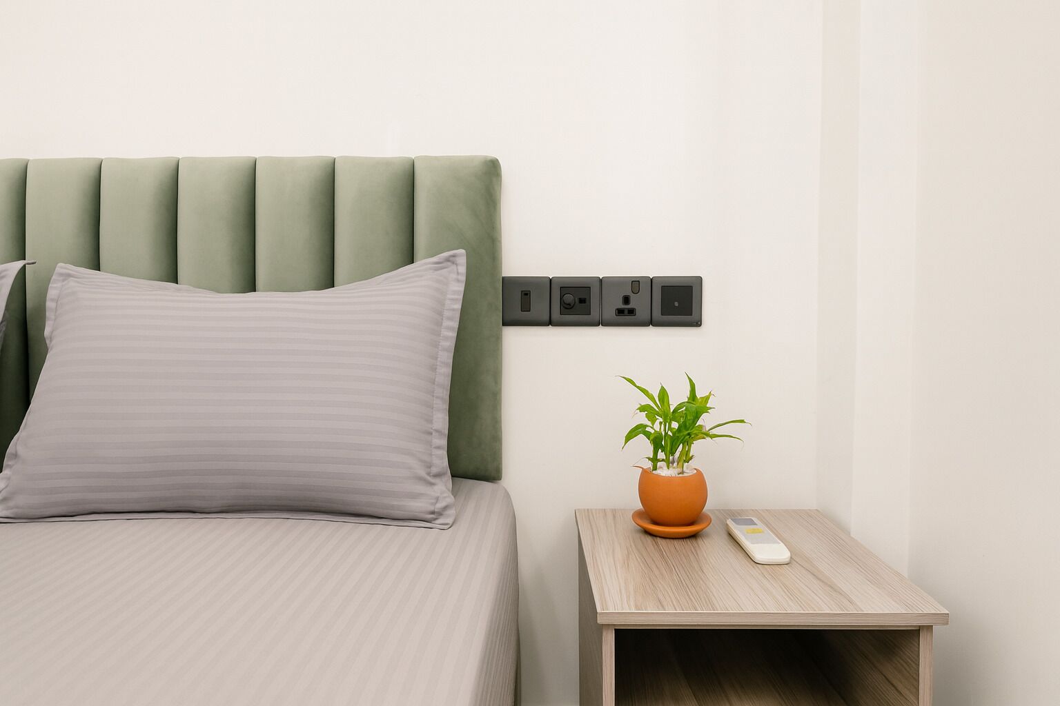 A bed with a green headboard, a grey pillow, a bedside table with a potted plant, and a remote control.