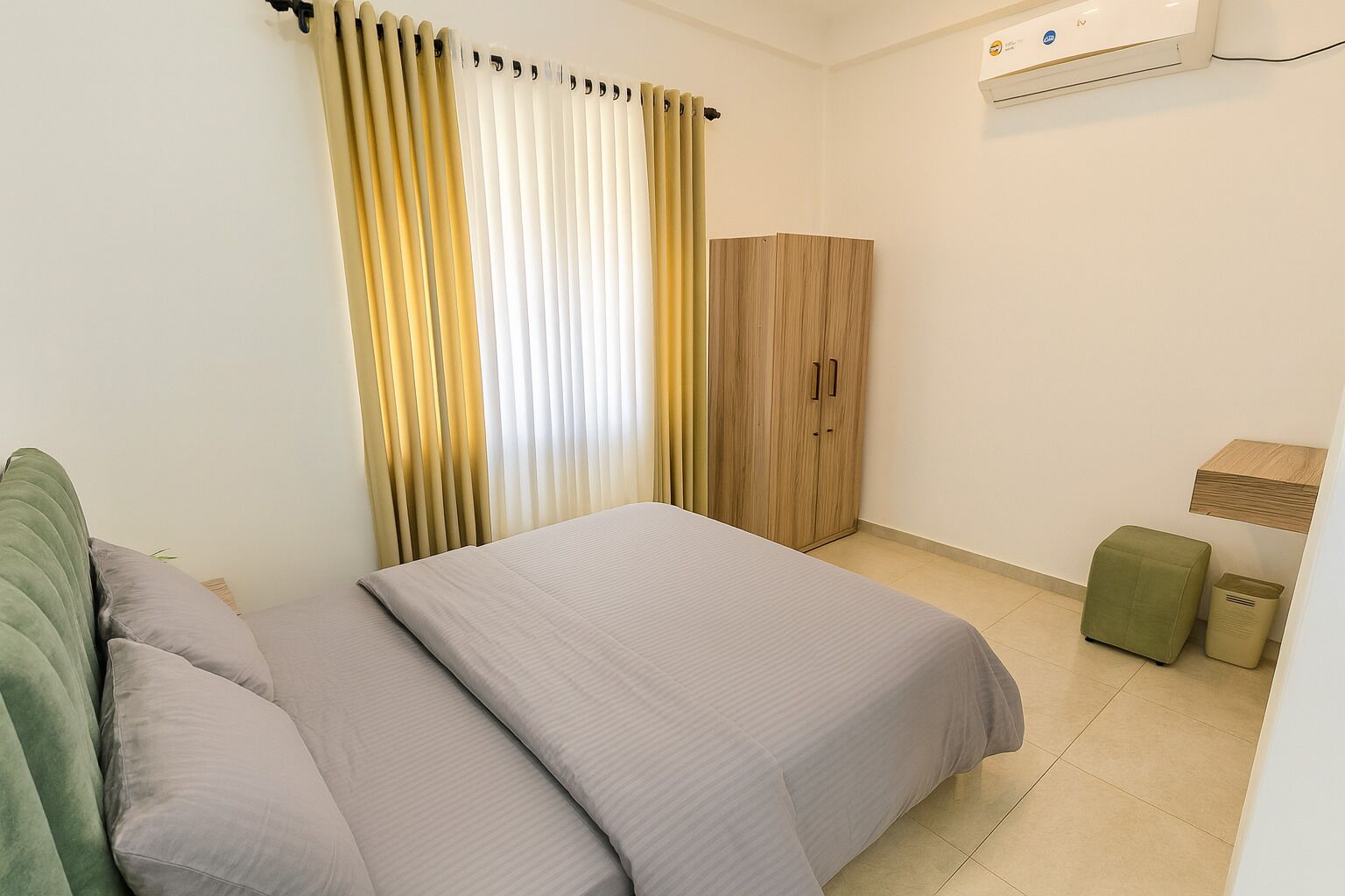 A bedroom with a bed, curtains, a wardrobe, a small table, and a trash bin.