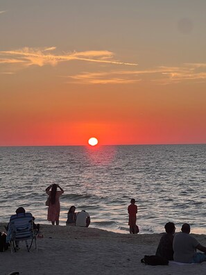 Une scène de plage avec des personnes assises et debout, un coucher de soleil et l’océan.