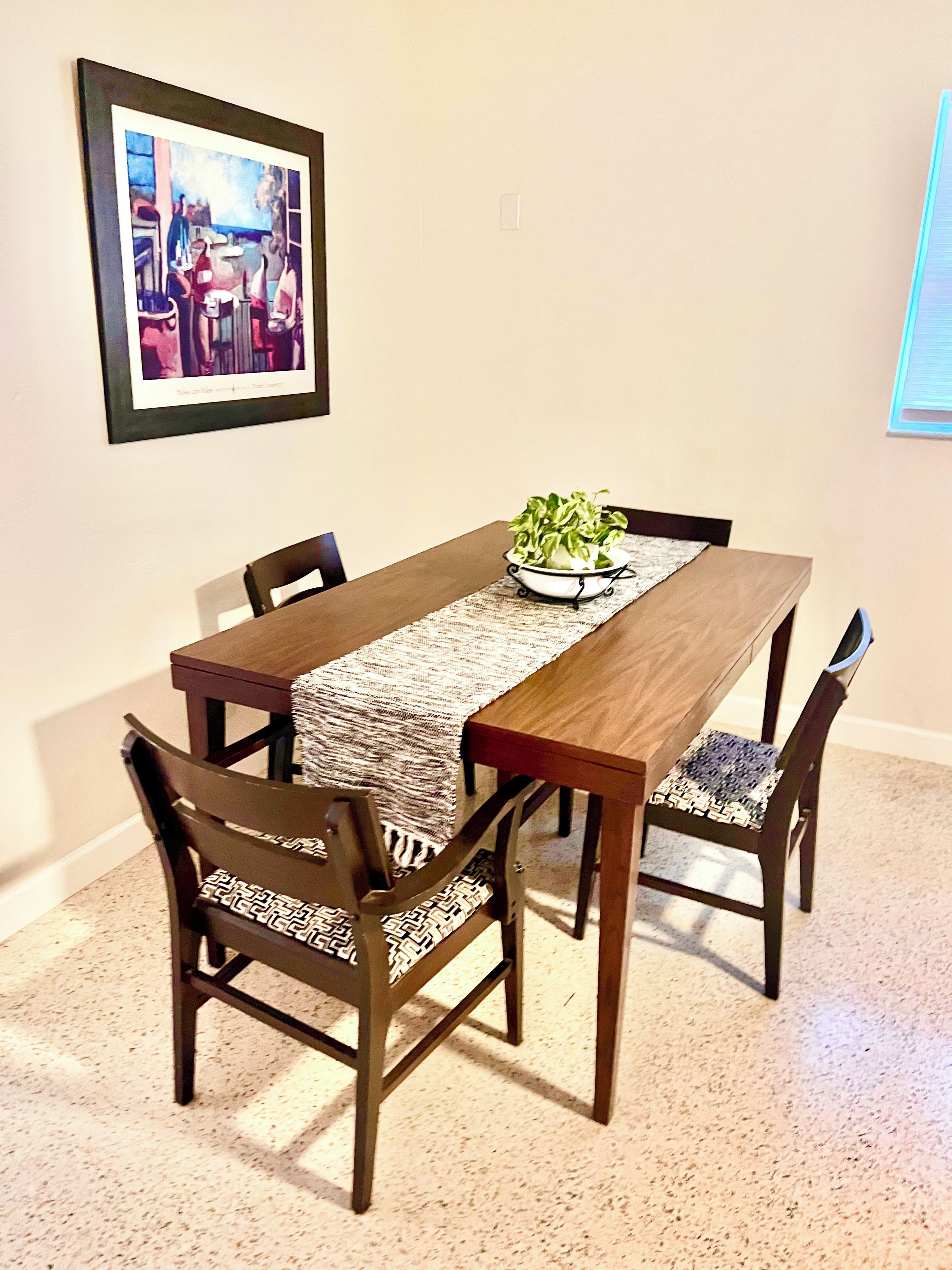 Un comedor con una mesa de madera, cuatro sillas, un camino de mesa tejido y una planta en maceta.