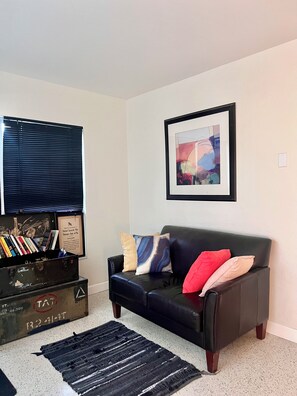 Un salon avec un canapé en cuir noir, un tableau abstrait encadré, une fenêtre avec des stores et une boîte de rangement contenant des livres et un certificat encadré.