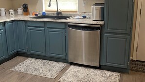 Uma cozinha com armários azuis, uma máquina de lavar louça de aço inoxidável e uma janela com cortina de renda.