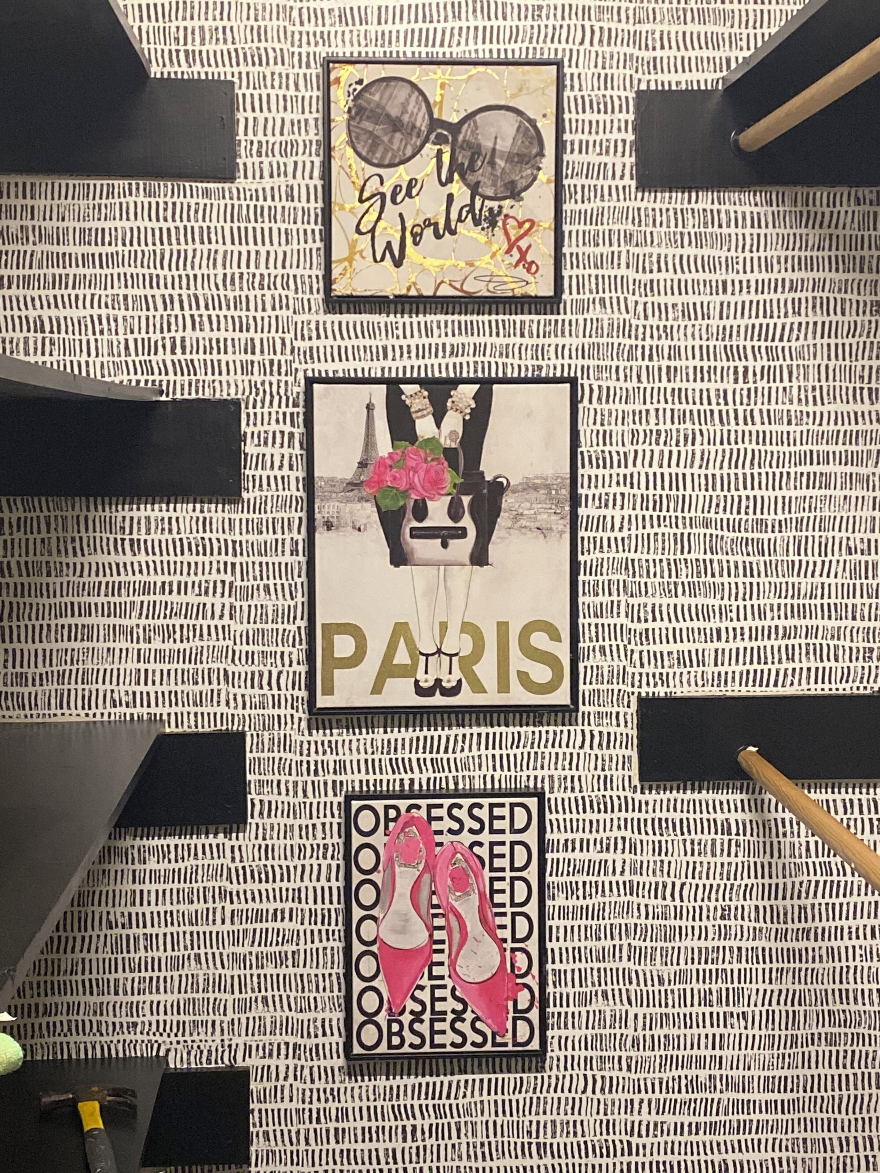 Un mur recouvert d’un papier peint à motifs et orné de trois objets encadrés : une paire de lunettes avec l’inscription « See the World », une illustration sur le thème de Paris représentant un sac à main, et une paire d’escarpins roses avec l’inscription « Obsessed ».
