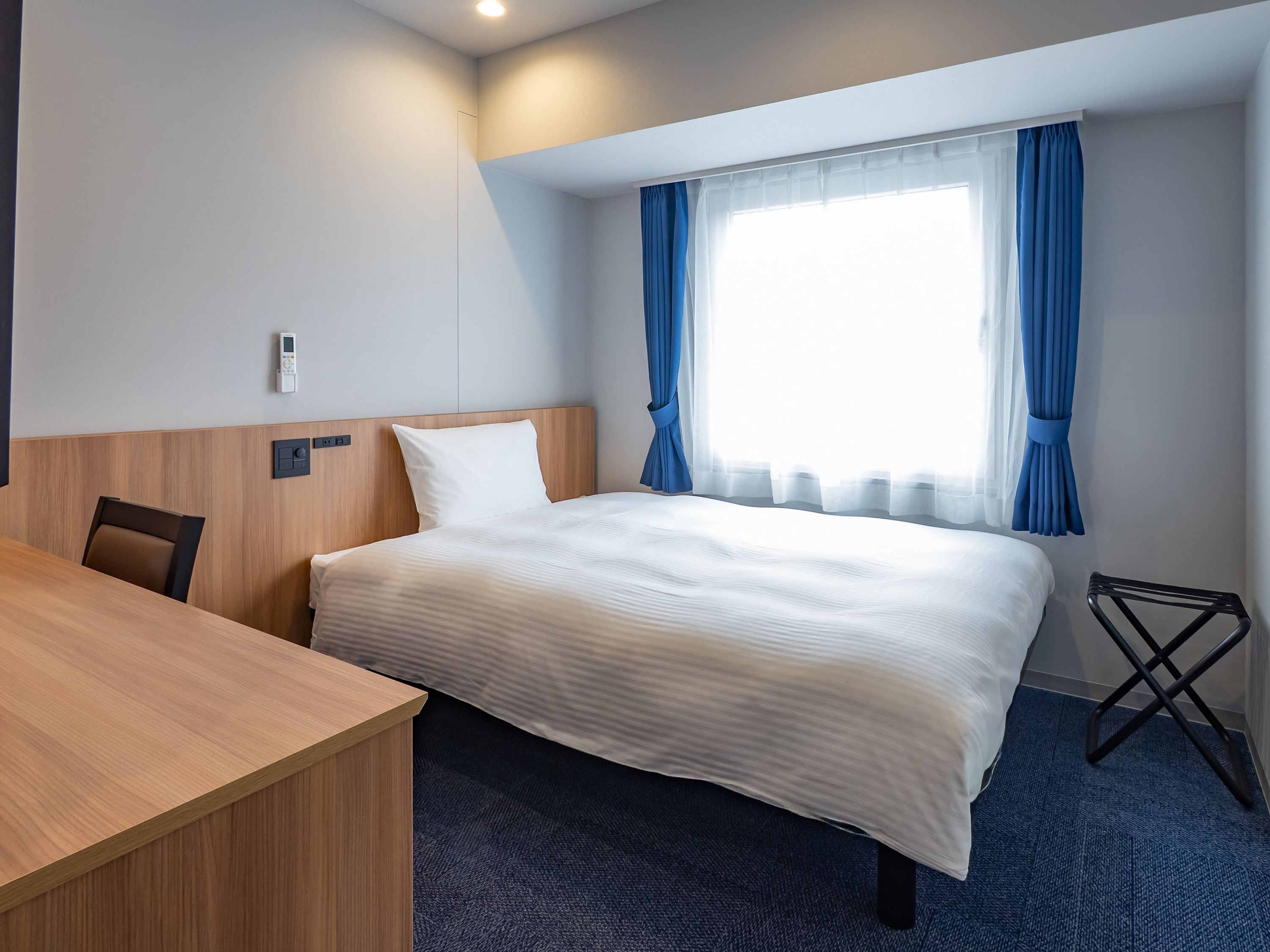 A hotel room with a bed, a wooden desk, a chair, a small table, and a window with curtains.
