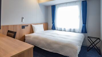 A hotel room with a bed, a wooden desk, a chair, a small table, and a window with curtains.