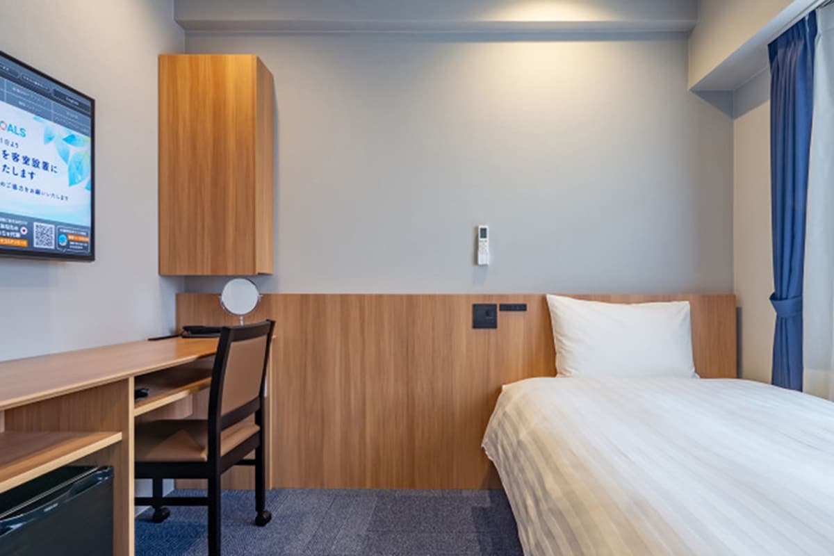 Quarto de hotel com cama, escrivaninha, cadeira, mesa pequena e televisão presa à parede.