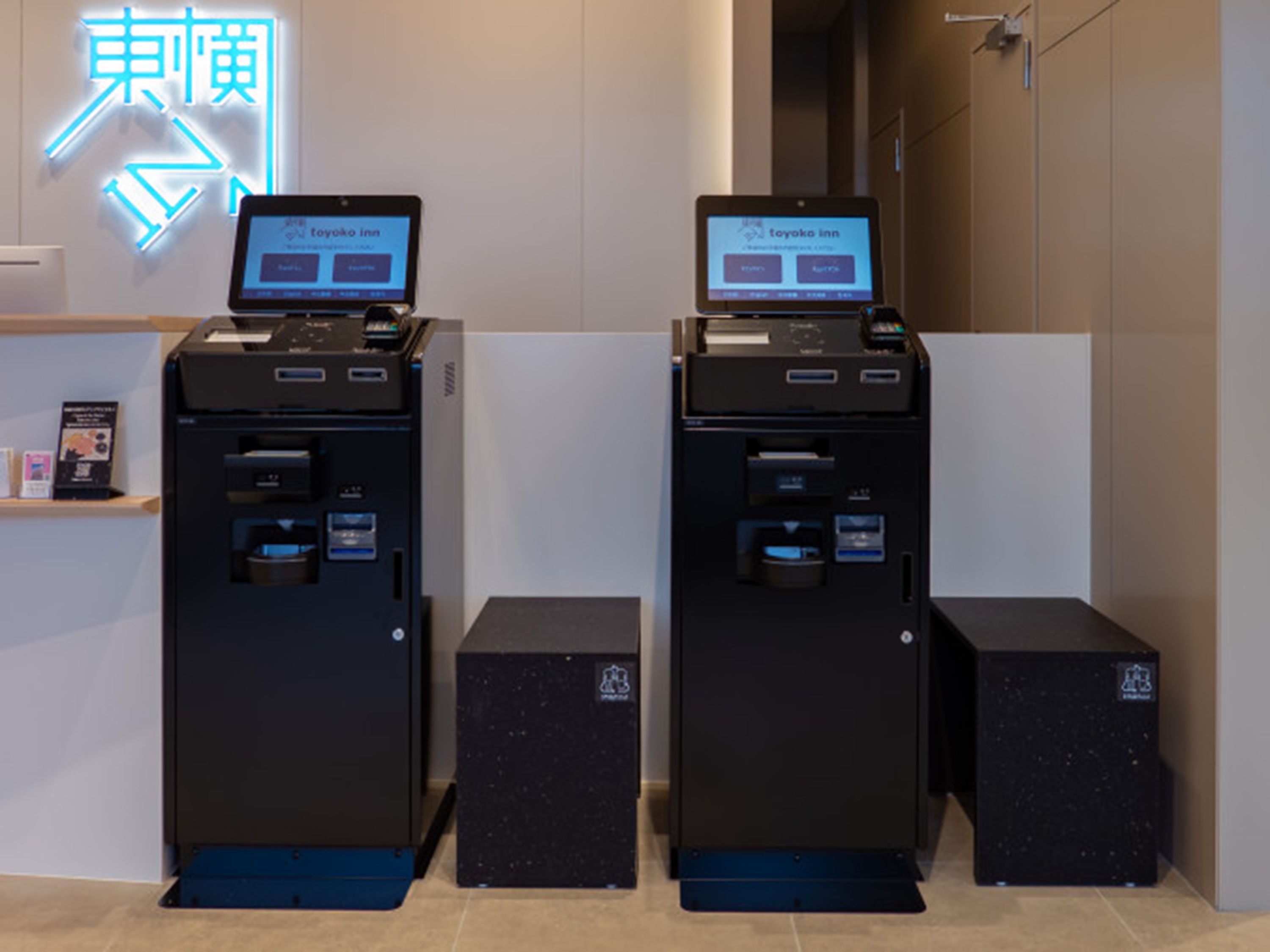 Two self-service kiosks with screens, a neon sign, and a small shelf with brochures.