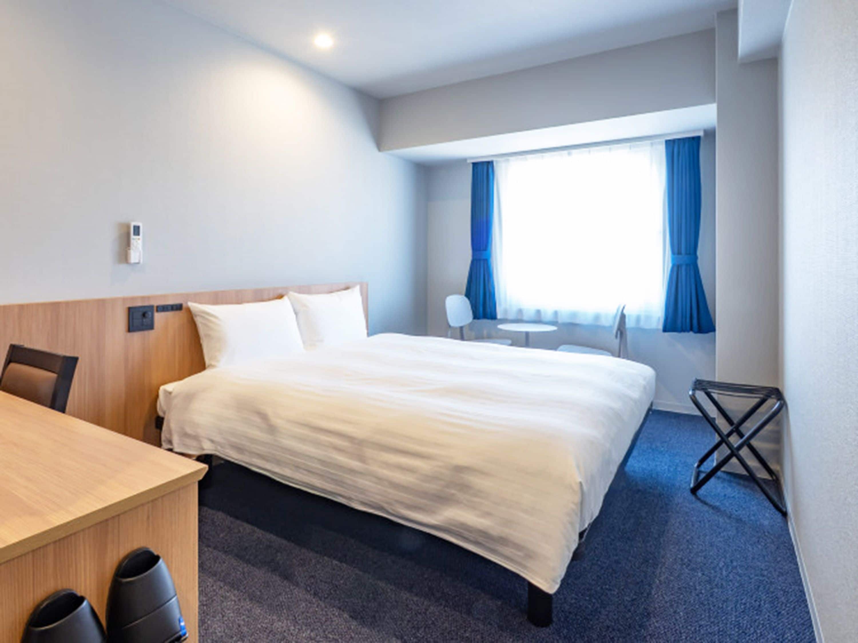 A hotel room with a bed, a desk, a chair, a small table, and a window with curtains.