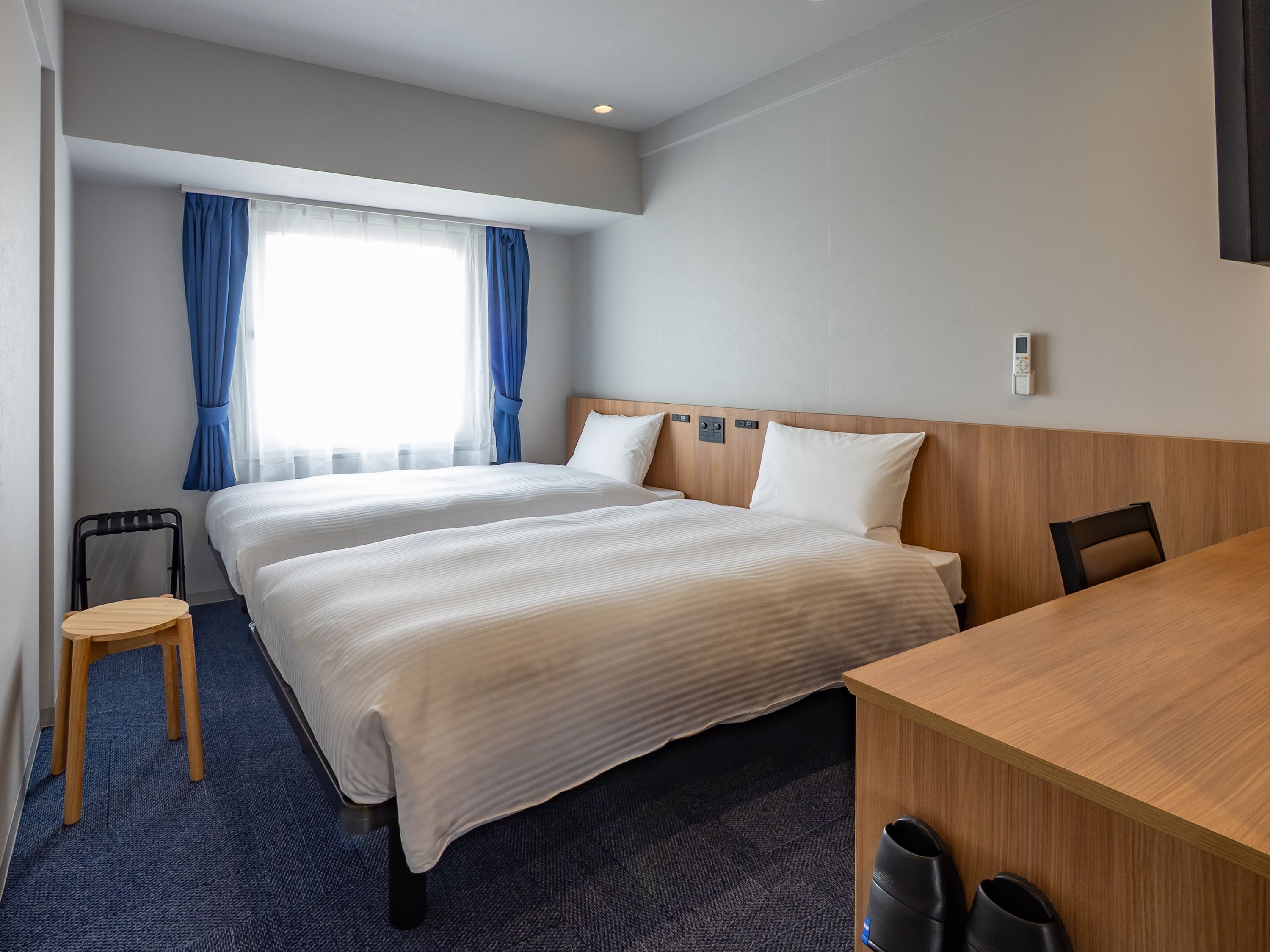 A hotel room with two beds, a wooden desk, a chair, and a window with curtains.