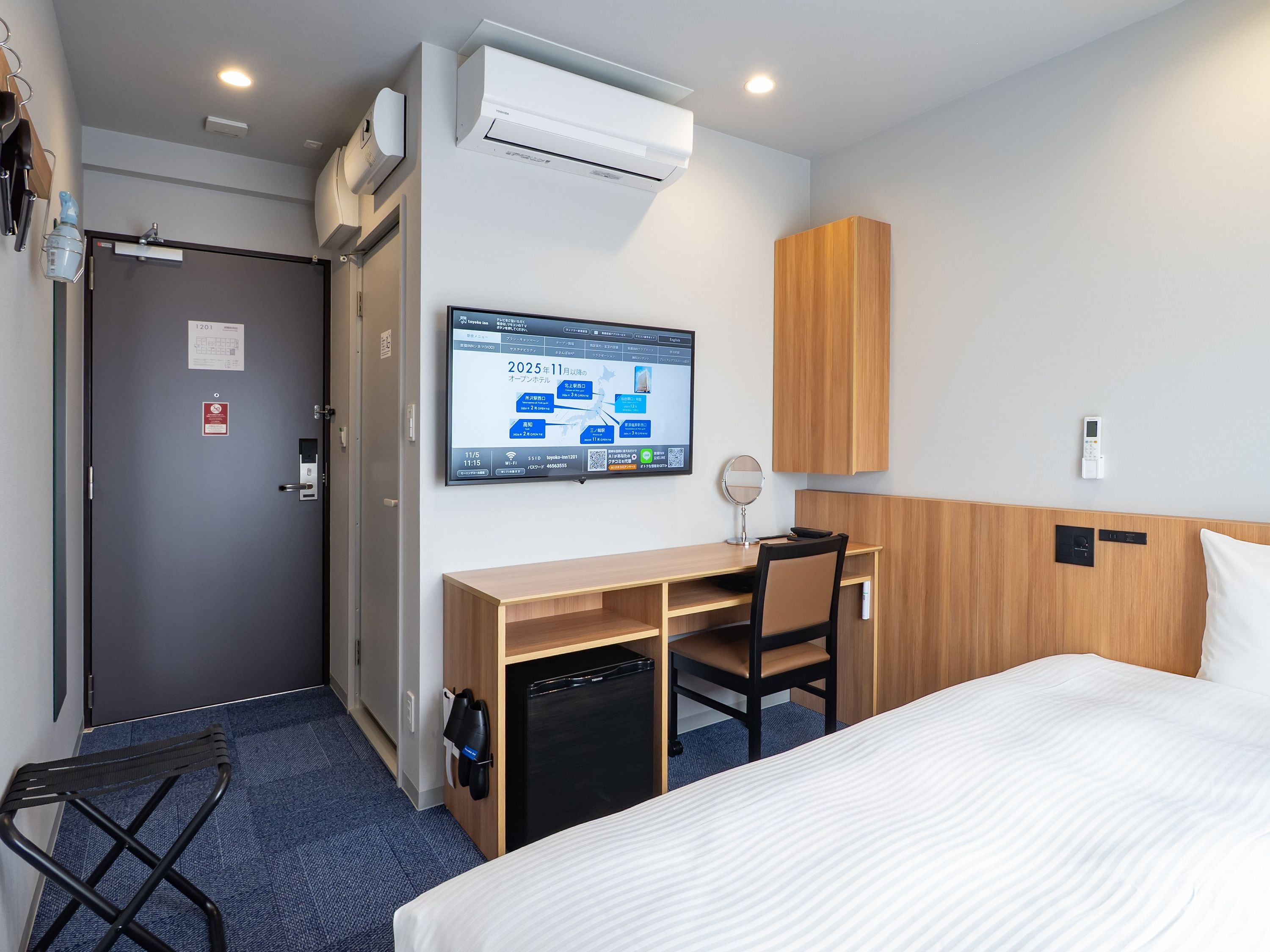 A hotel room with a bed, a desk, a chair, a TV, an air conditioner, and a small refrigerator.