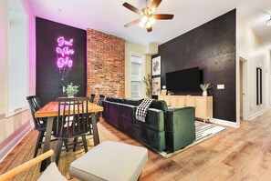 A modern living room with a dining area, a black brick accent wall, a neon sign, a ceiling fan, and a flat-screen TV. - Netflix+BBQ+Free Parking Near Shops in East Liberty (Pittsburgh)