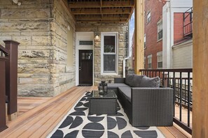 A covered porch with stone walls, a wooden deck, a black and white patterned rug, and outdoor furniture. - Netflix+BBQ+Free Parking Near Shops in East Liberty (Pittsburgh)