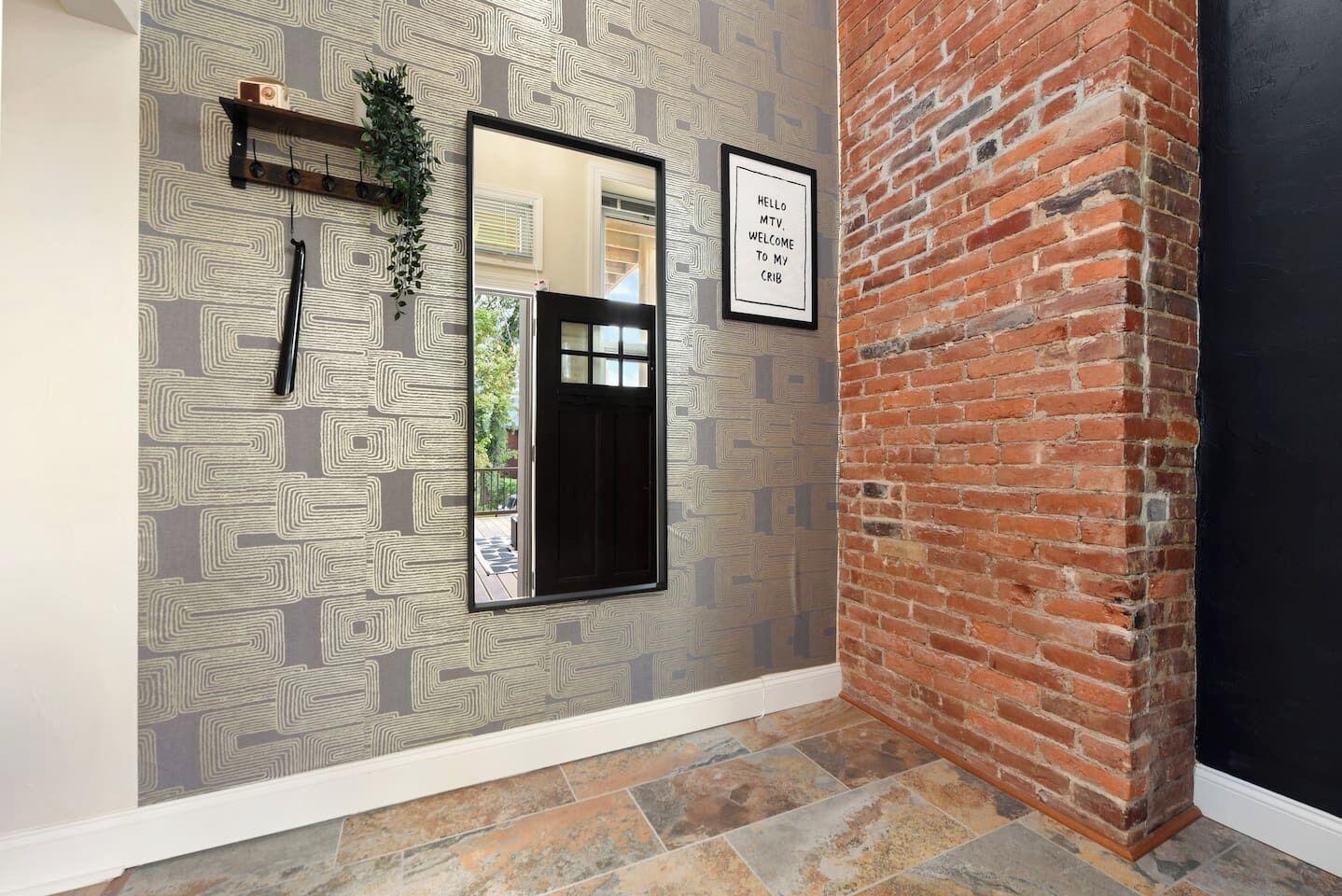 A hallway with patterned wallpaper, a black-framed mirror, a welcome sign, and a brick wall.