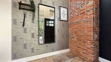 A hallway with patterned wallpaper, a black-framed mirror, a welcome sign, and a brick wall.