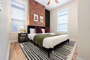 Une chambre avec un mur en briques, un lit avec des oreillers blancs et bordeaux, un plaid vert et un tapis rayé noir et blanc.