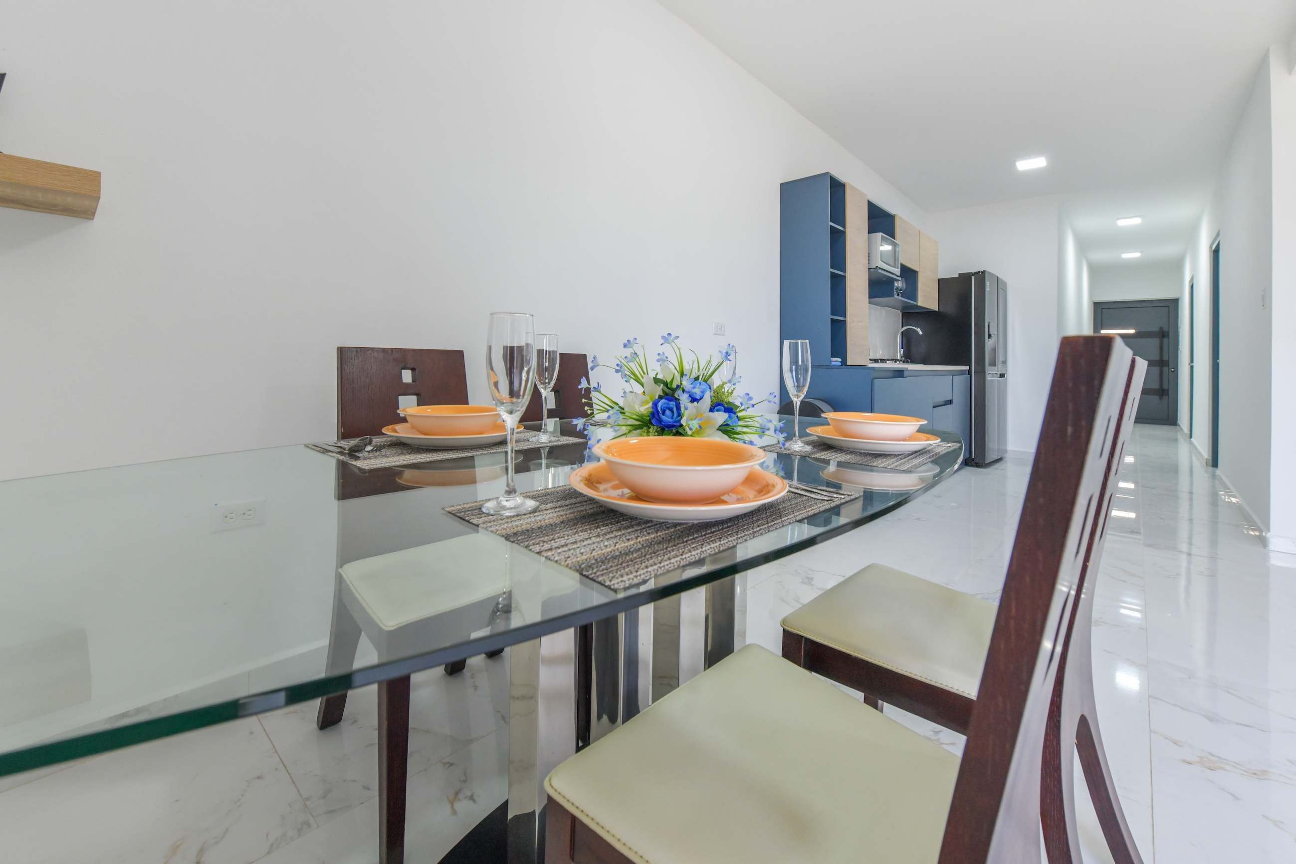 Uma área de jantar moderna com uma mesa de vidro, cadeiras brancas e uma cozinha ao fundo.