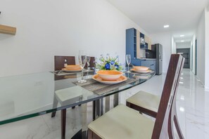 A modern dining area with a glass table, white chairs, and a kitchen in the background. - Sleek 2BR Retreat, Tanki Leendert (Tanki Leendert)