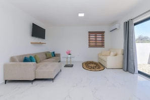 A modern living room with a sofa, a coffee table, a TV mounted on the wall, and a window with blinds. - Sleek 2BR Retreat, Tanki Leendert (Tanki Leendert)