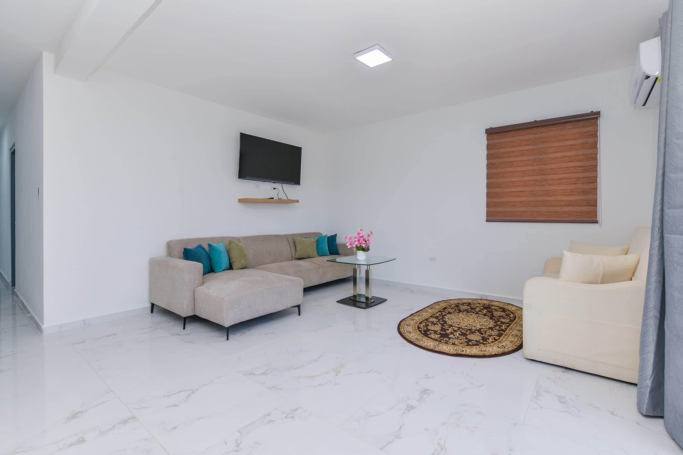 A modern living room with a sectional sofa, a small round table, a flat-screen TV mounted on the wall, and a window with blinds.