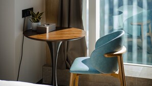 Ett hotellrum med en säng, ett litet bord med en växt, en stol och utsikt över en stadssiluett genom ett stort fönster.