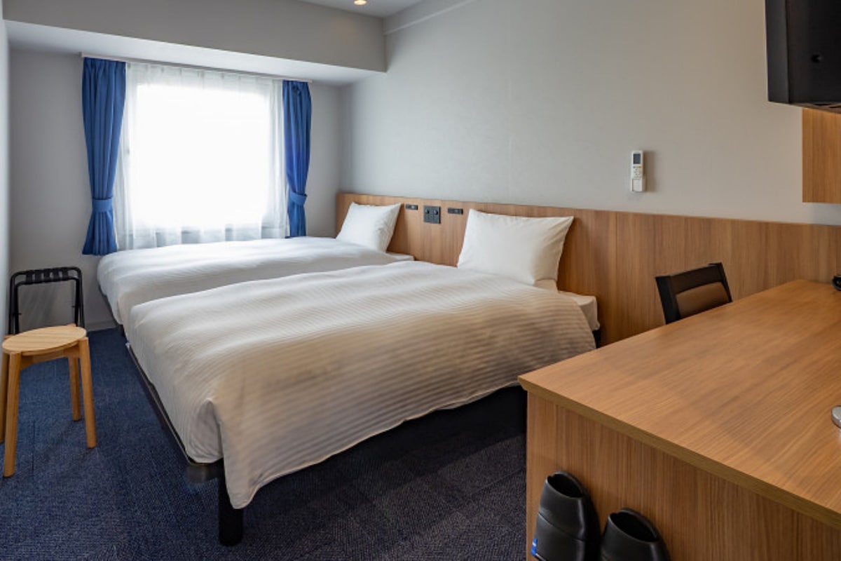 Habitación de hotel con dos camas, un escritorio de madera, una silla, un televisor y un ventanal con cortinas.