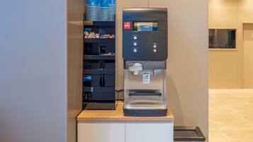 A water dispenser on a counter with a coffee machine beside it, and a stack of blue cups on the wall.