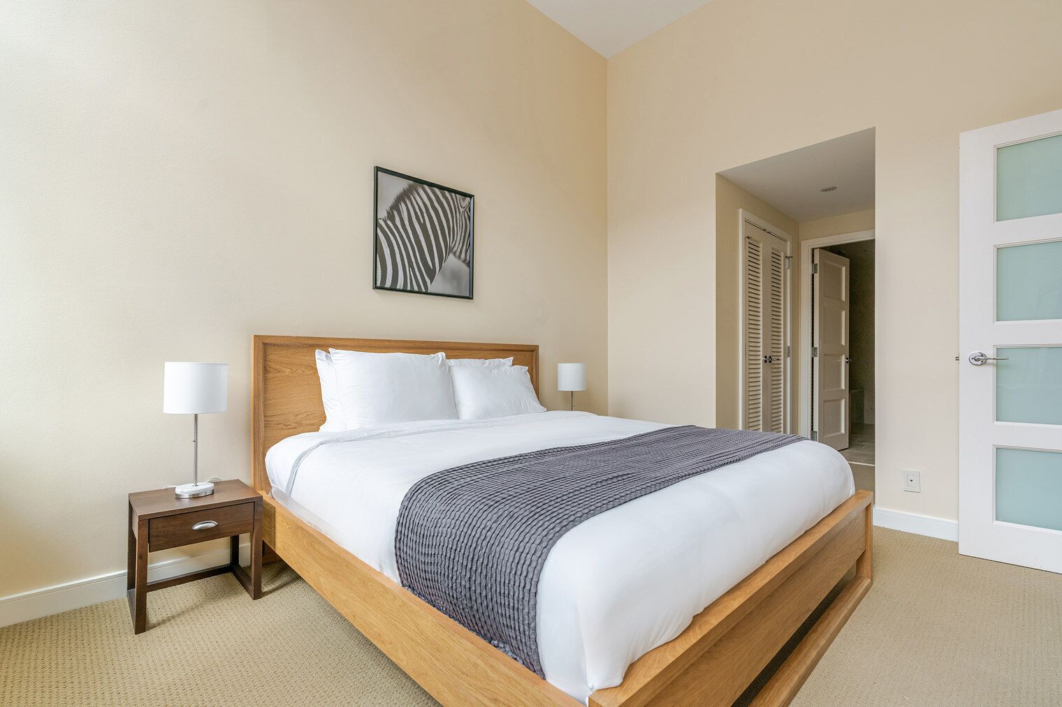 A hotel room with a wooden bed, white bedding, a gray throw, two bedside lamps, a framed artwork on the wall, and a door with frosted glass panels.