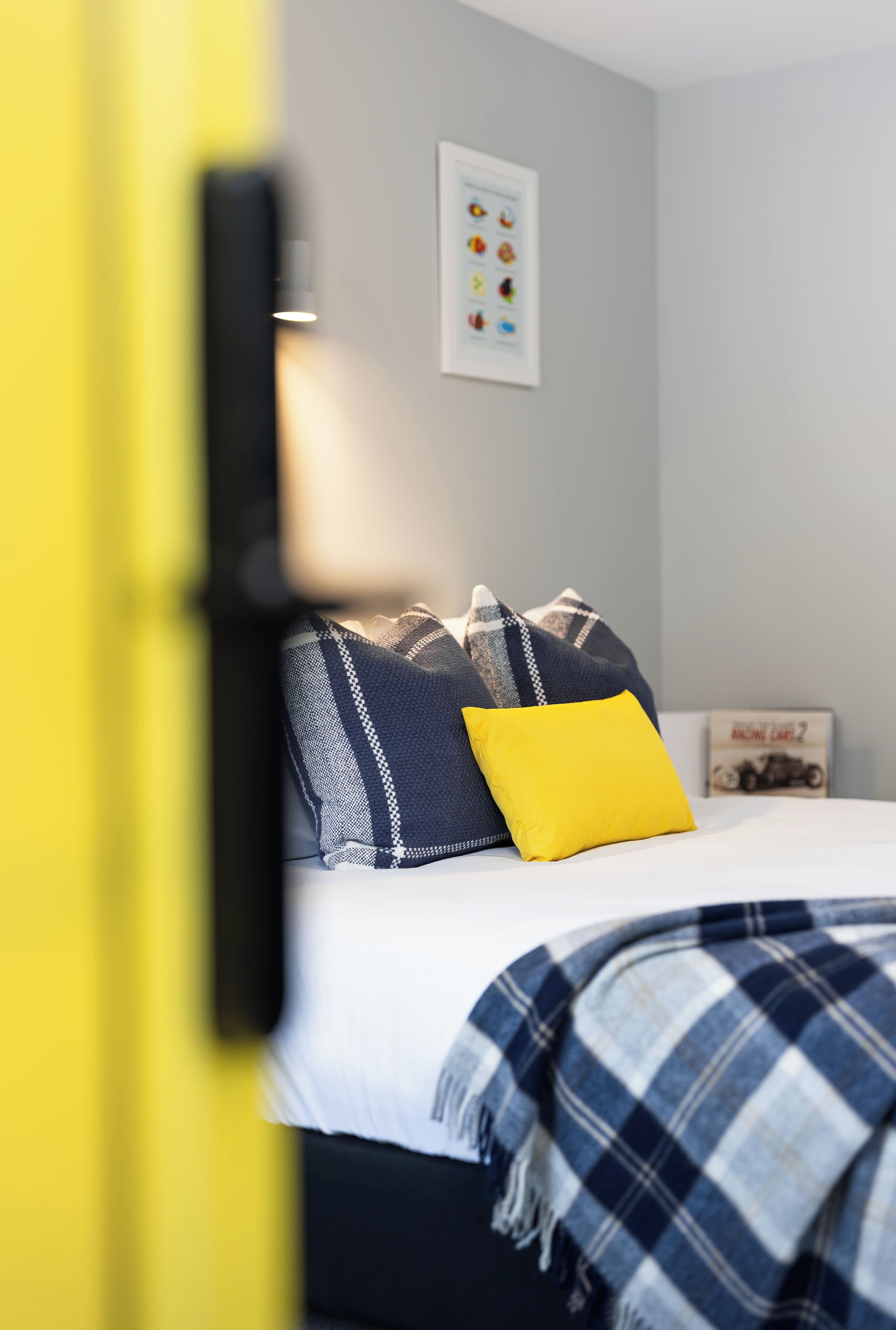Uma cama com colchão branco, manta azul e branca xadrez e travesseiro amarelo.