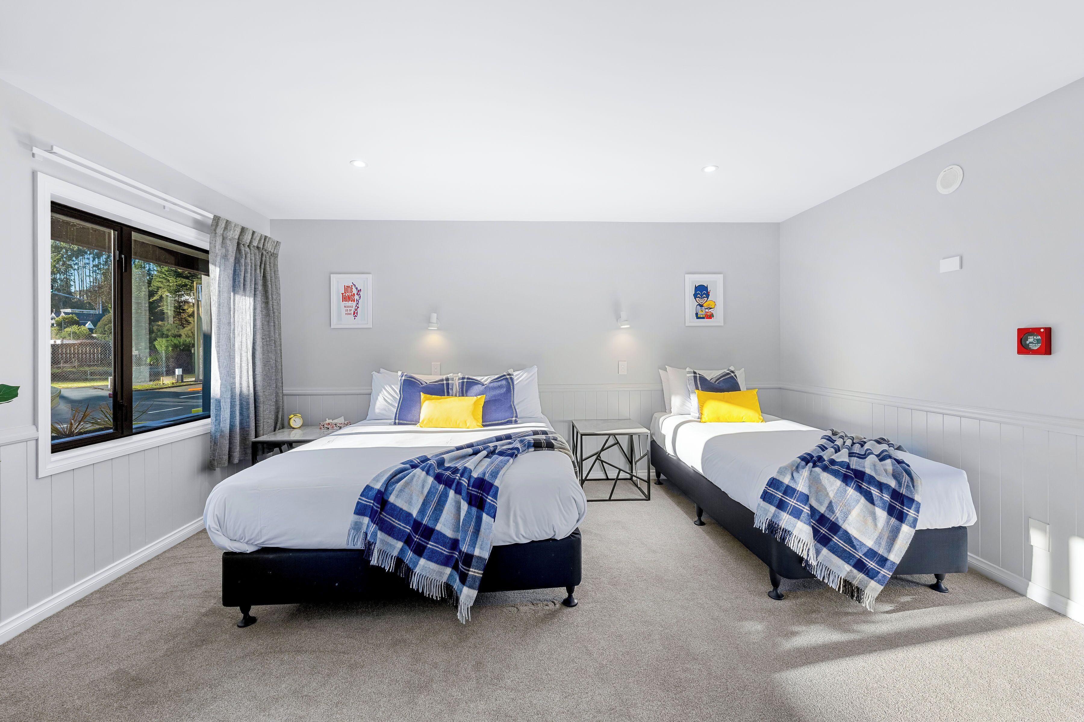 A hotel room with two beds, a window with curtains, a small table, and a wall-mounted light.