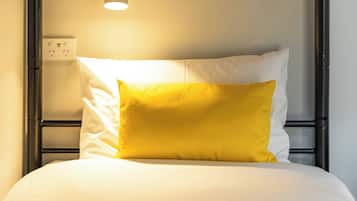 Ein Bett mit weißer Matratze und einem gelben Kissen, ein Nachttisch mit Lampe sowie eine Steckdose an der Wand.