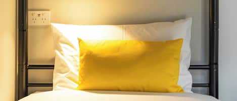 A bed with a white mattress and a single yellow pillow, a bedside table with a lamp, and a power outlet on the wall.