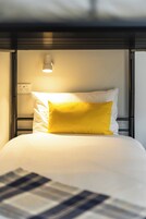 Una cama con colchón blanco y una sola almohada amarilla, una mesita de noche con lámpara, y un enchufe en la pared.