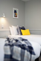 Un dormitorio con cama, almohadas, una colcha, una lámpara de pared y un cuadro enmarcado.