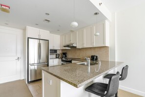 A modern kitchen with white cabinets, stainless steel appliances, a granite countertop, and a bar area with two chairs. - Blueground | Penn Quarter, pool, nr National Gallery (Washington, D.C.)