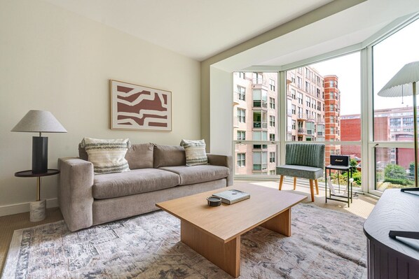 A modern living room with a sofa, coffee table, and a lamp. There is a painting on the wall and a view of buildings outside the window. - Blueground | Penn Quarter, pool, nr National Gallery (Washington, D.C.)