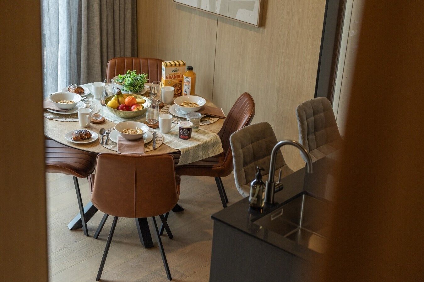 A dining area with a round table set for four, surrounded by brown upholstered chairs. A bottle of granola, a bowl of fruit, and various breakfast items are visible on the table.