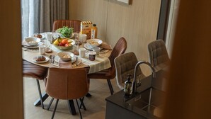 Un'area pranzo con un tavolo rotondo apparecchiato per quattro persone, circondato da sedie imbottite marroni. Sul tavolo si vedono una bottiglia di granola, una ciotola di frutta e diversi articoli per la colazione.
