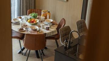 A dining area with a round table set for four, surrounded by brown upholstered chairs. A bottle of granola, a bowl of fruit, and various breakfast items are visible on the table.