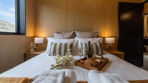 Una camera d'hotel con un letto grande, due comodini, un tavolino da notte, una testiera in legno e una finestra con vista su un paesaggio desertico.