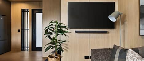 A modern living room with a flat-screen TV mounted on the wall, a sofa with patterned cushions, a floor lamp, and a potted plant.