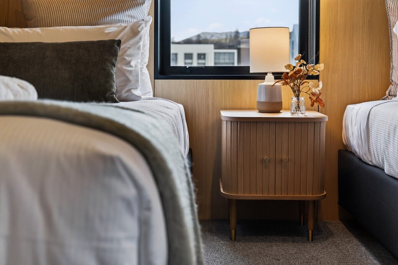 A hotel room with a bed, a bedside table with a lamp, and a window with a view of buildings.
