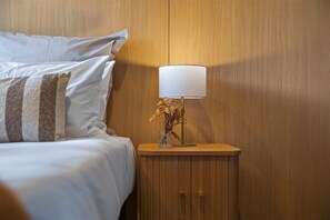 A neatly made bed with white bedding and striped pillows, a bedside table with a lamp and a vase of dried flowers, and wooden paneling on the wall.