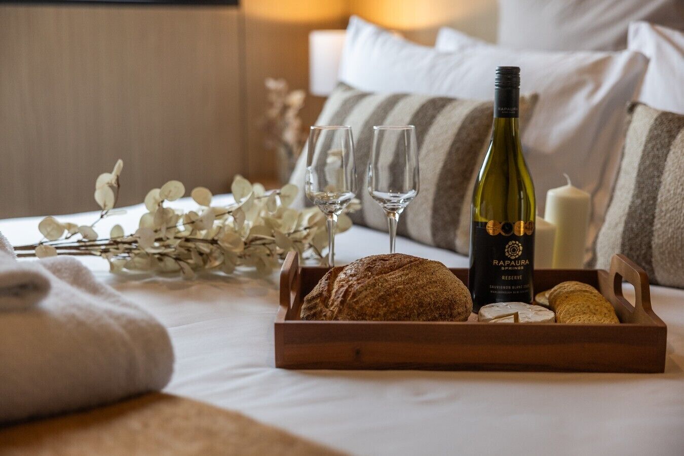 A bed with a wooden tray containing a loaf of bread, a bottle of wine, and two glasses.