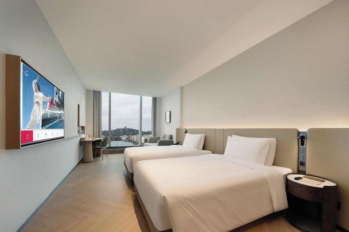 Quarto de hotel moderno com duas camas, uma escrivaninha e uma janela grande com vista para a cidade.