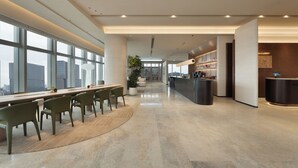 Un moderno vestíbulo de hotel con una larga recepción, área de descanso con sillas, una gran planta en maceta y vistas de la ciudad a través de amplias ventanas.