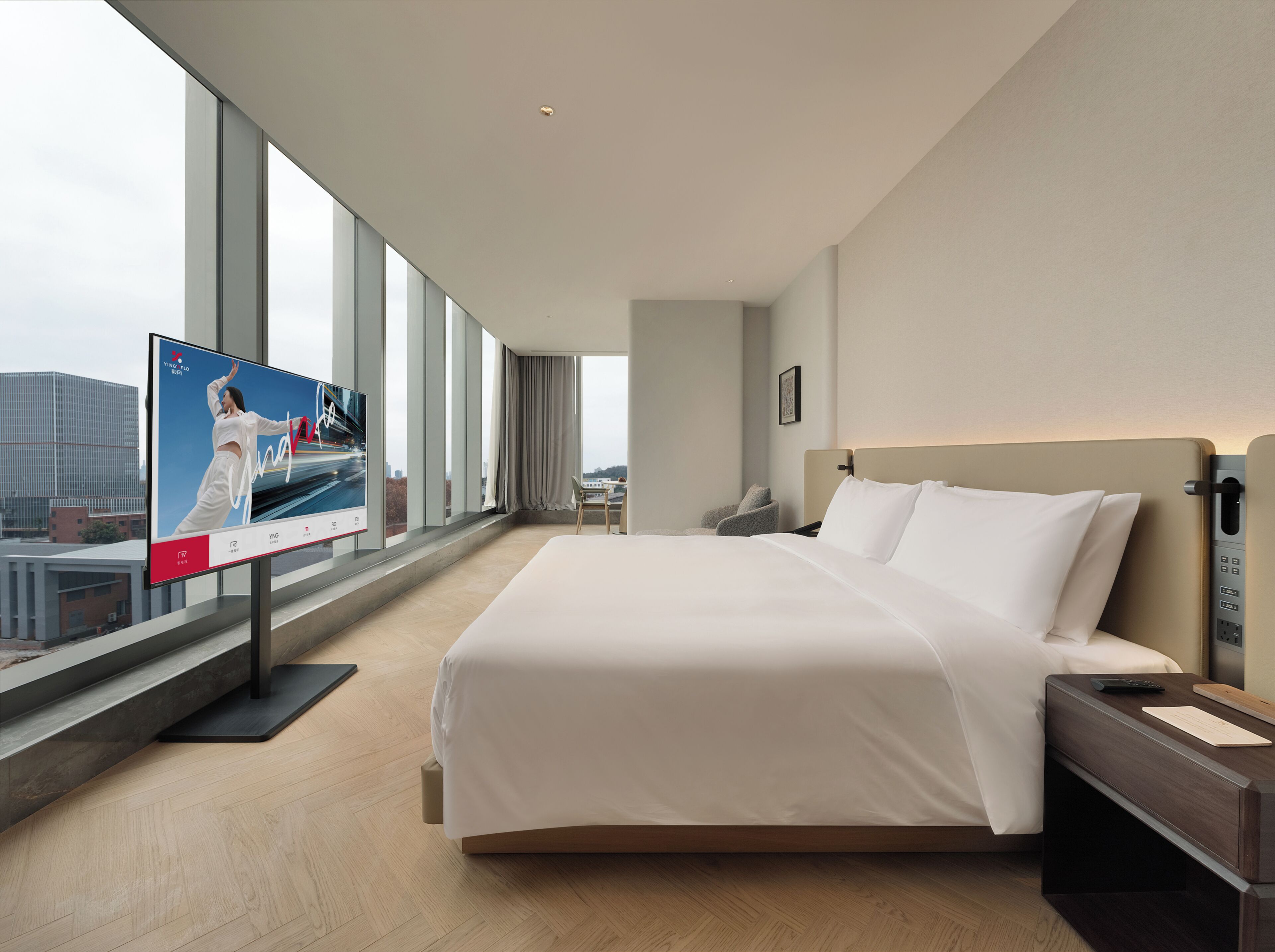 Habitación de hotel con una cama grande, un escritorio, una silla, un televisor y vistas a la ciudad a través de amplios ventanales.